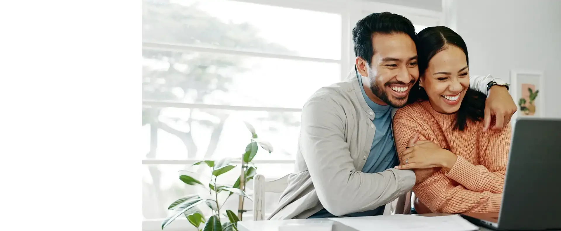 A couple sits close together at a table with laptops and a plant, sharing a cozy moment in a bright, modern workspace.